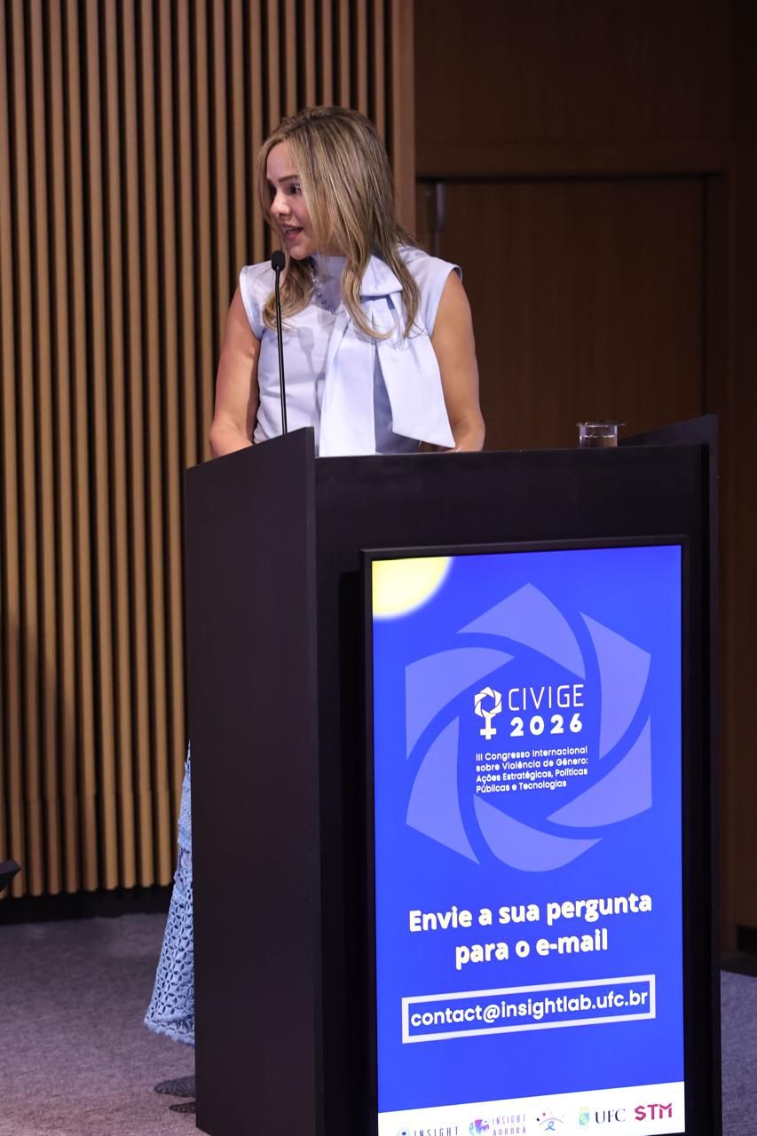 A Dr.ª Amini Haddad, Juíza Auxiliar e Coordenadora do Observatório Pró-Equidade do STM, discursa atrás de um púlpito preto com uma tela de LED que exibe o logotipo "CIVIGE 2026". Ela veste uma roupa azul-marinho e fala ao microfone.