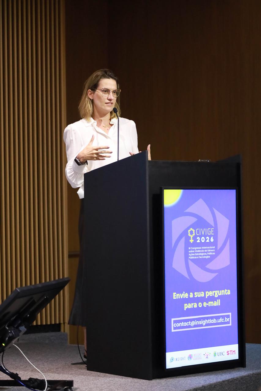 A Dr.ª Carla Pinheiro Bessa Von Bentzen Rodrigues, Procuradora do Estado de Goiás, discursa atrás de um púlpito preto. O monitor do púlpito exibe o logotipo 'CIVIGE 2026' e o e-mail 'contact@insightlab.ufc.br' para perguntas. Ela veste camisa branca e saia longa preta com óculos de armação clara contra um fundo de madeira ripada.