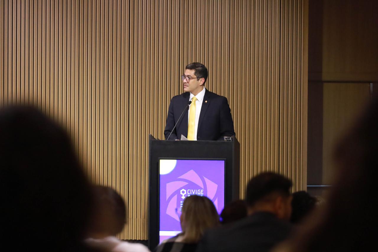 A foto mostra o Dr. Flávio Henrique Albuquerque de Freitas, Juiz Auxiliar da Presidência, discursando atrás de um púlpito preto com uma tela que exibe o logotipo roxo e branco "CIVIGE". Ele veste terno preto, camisa branca e uma gravata amarela brilhante, falando ao microfone contra um fundo de parede de madeira ripada, com silhuetas desfocadas da plateia em primeiro plano.