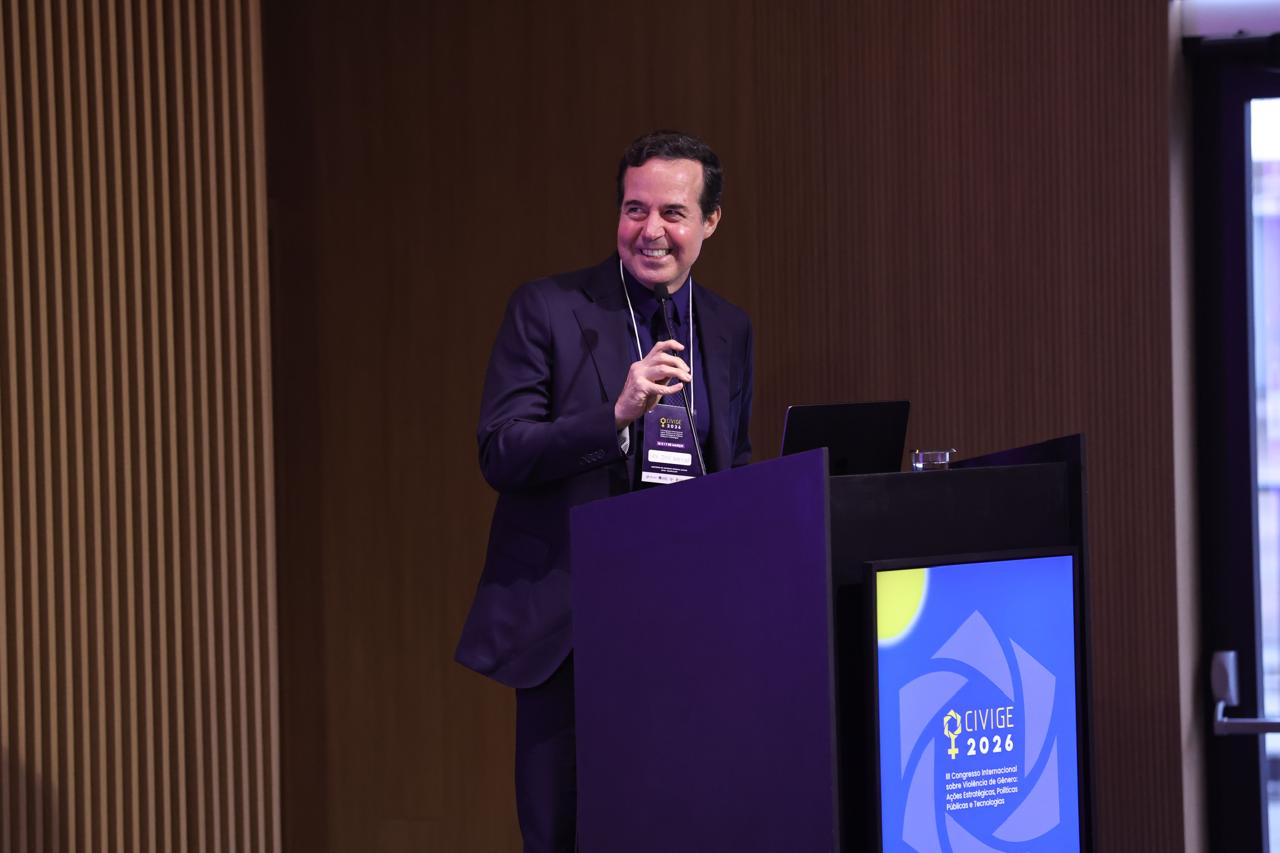 O Prof. José Macêdo sorri enquanto fala atrás de um púlpito preto durante o III CIVIGE 2026. Ele veste terno escuro e usa um crachá de identificação. O monitor acoplado ao púlpito exibe o logotipo do evento, com um fundo de ripas de madeira ao lado.