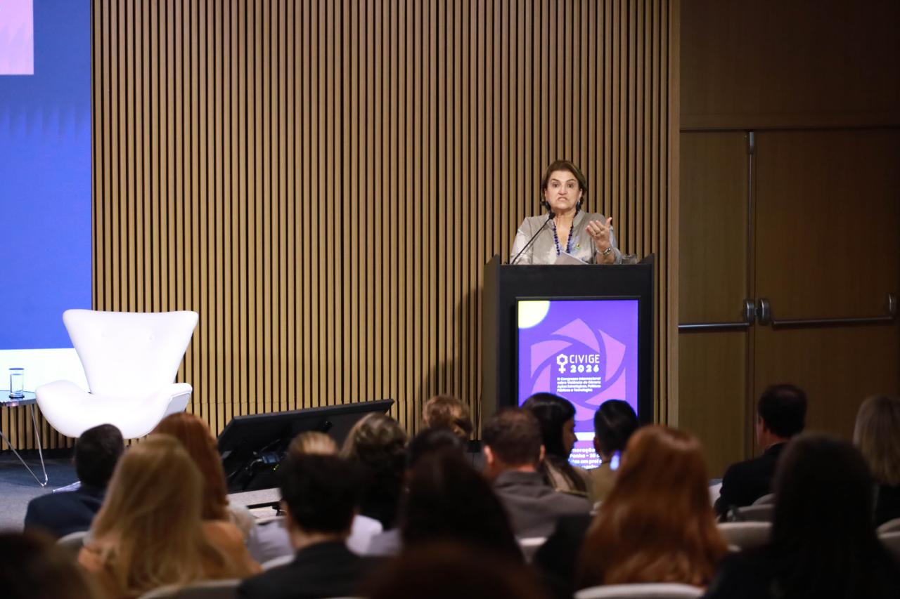 A Ministra das Mulheres, Márcia Lopes, discursa em um palco de auditório, atrás de um púlpito com a tela 'CIVIGE 2026'. Uma plateia está sentada em primeiro plano e a parede de fundo tem ripas de madeira.