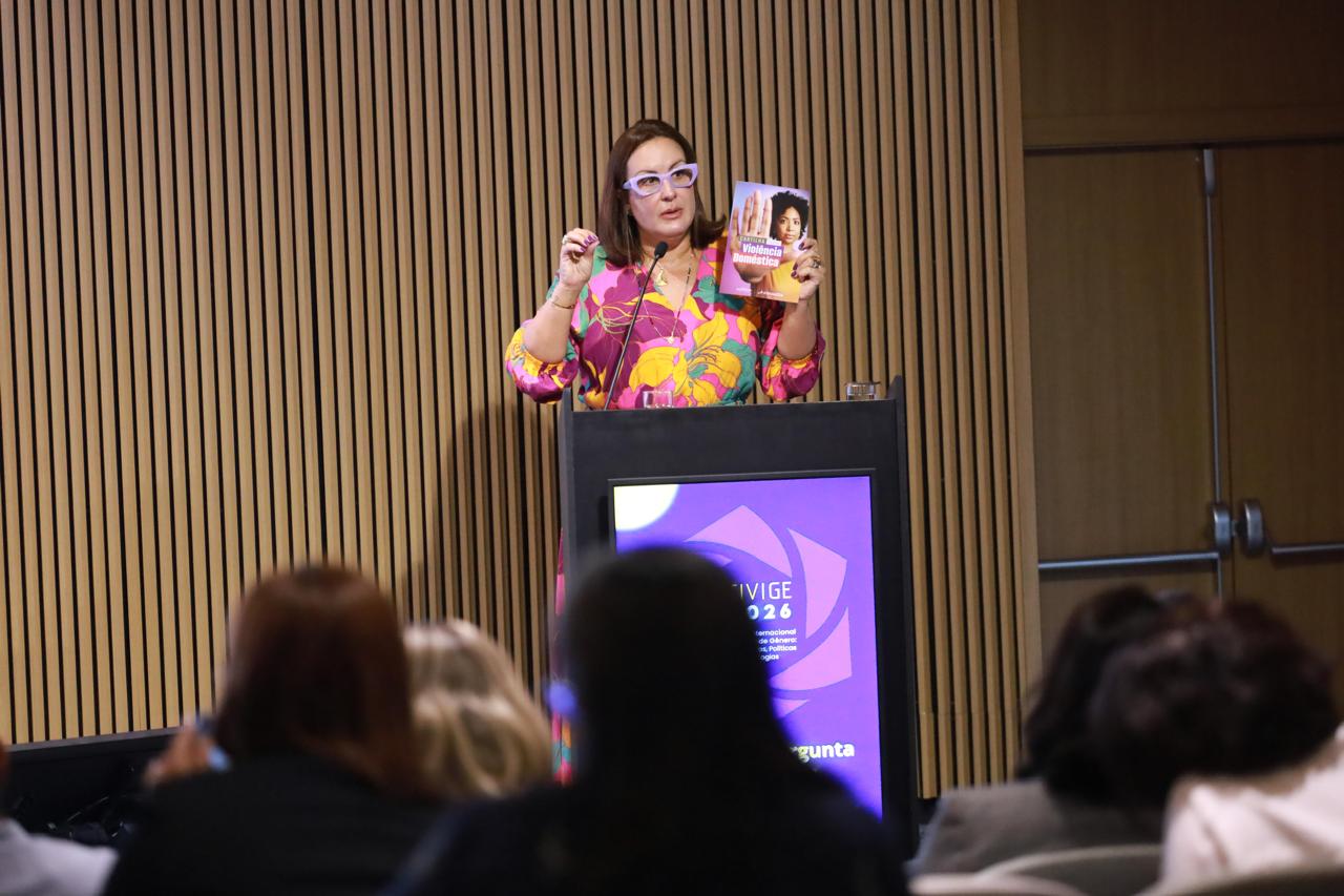 A Dr.ª Patrícia Sant’Anna, Juíza do Trabalho do TRT 12, discursa em um púlpito preto durante o CIVIGE 2026. Ela usa óculos de armação lilás e um vestido colorido com estampa floral, segurando uma cartilha sobre Violência Doméstica para a plateia. Ao fundo, uma parede de ripas de madeira e, no púlpito, um monitor exibe o logotipo do evento.