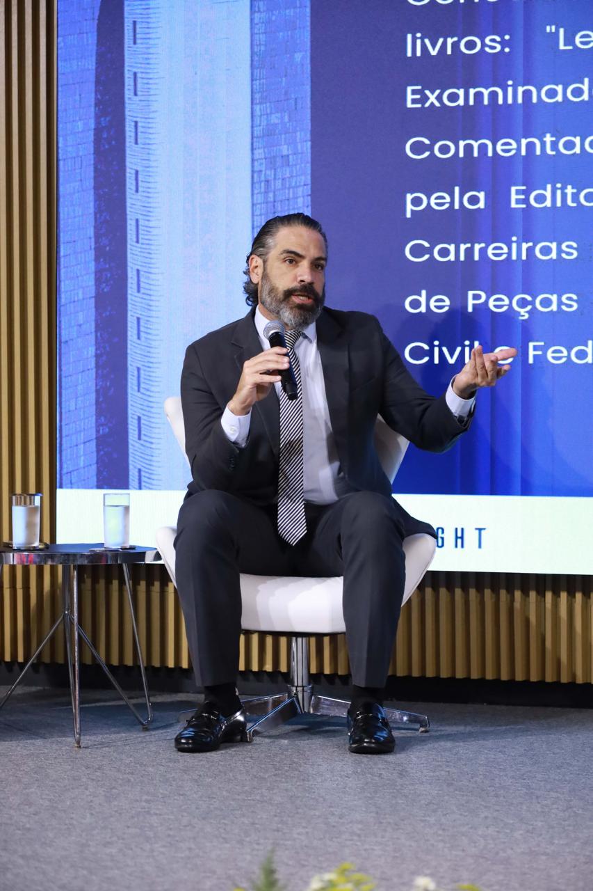 Dr. Sérgio Bautzer, Delegado da Polícia Civil do Distrito Federal (PCDF), sentado em uma poltrona branca moderna em um palco. Ele está vestido com um terno cinza-chumbo, camisa social branca e uma gravata com listras diagonais, segurando um microfone na mão direita enquanto fala e gesticula com a mão esquerda. Atrás dele, há uma grande tela de LED. Ao lado esquerdo, há uma pequena mesa auxiliar com dois copos de água. O palco tem piso cinza.