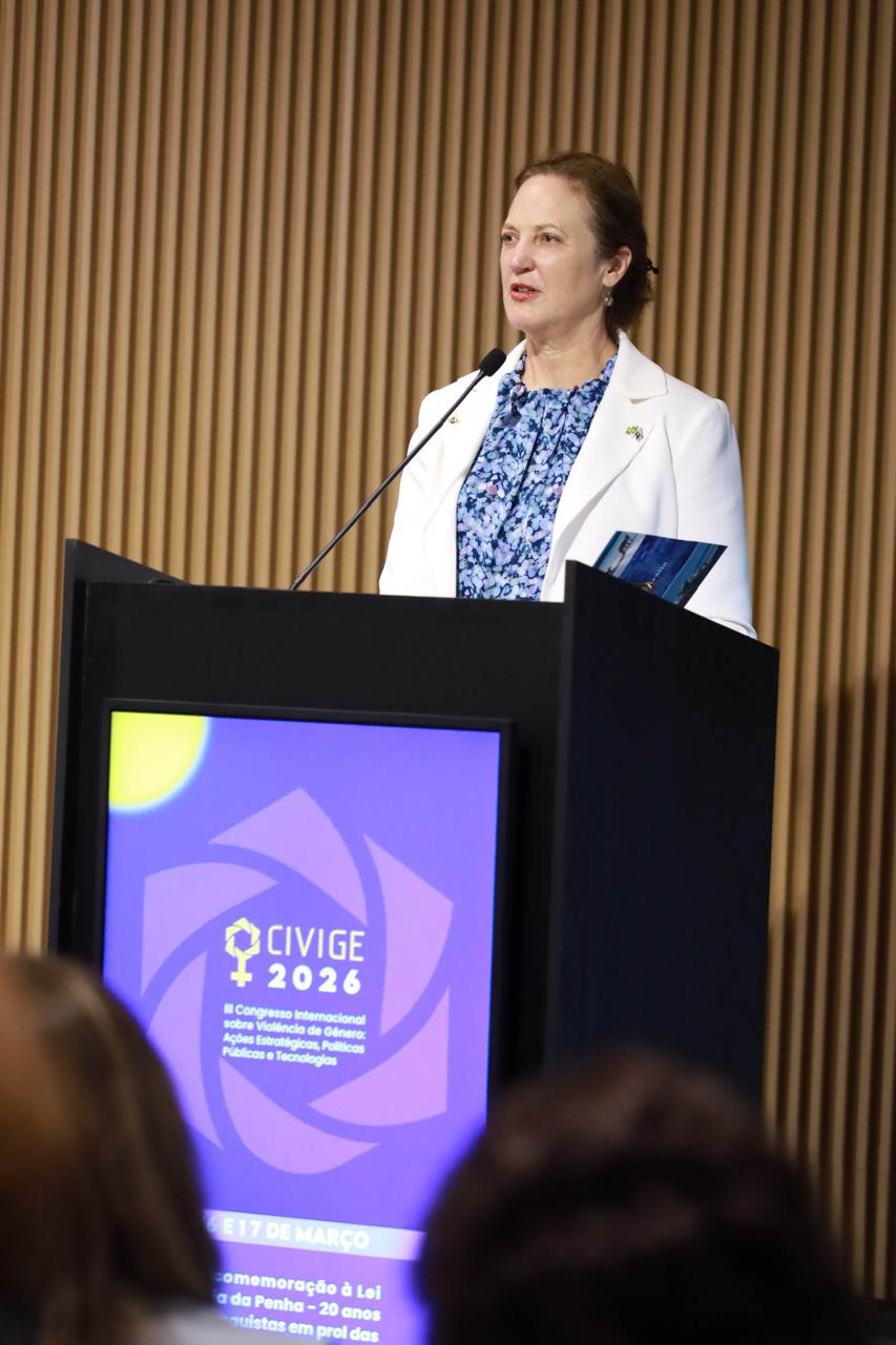 A Embaixadora da Austrália, Sophie Davies, discursa em um púlpito preto. O monitor integrado ao púlpito exibe o logotipo "CIVIGE 2026". Ela veste um blazer branco e uma blusa estampada contra um fundo de madeira ripada.