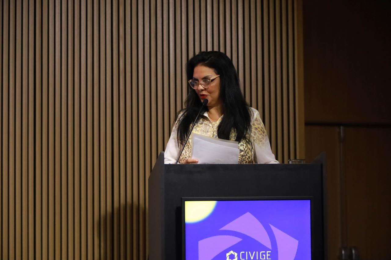 A Dr.ª Tânia Regina Silva Reckziegel, Desembargadora e Juíza Auxiliar da Presidência, discursa em pé atrás de um púlpito preto. Ela está segurando e olhando para folhas de papel em suas mãos. Ela veste uma camisa de manga longa estampada, com detalhes em branco e marrom, e óculos de armação clara. O monitor do púlpito exibe o logotipo do evento CIVIGE sobre um fundo roxo e azul. O fundo da imagem é uma parede de ripas de madeira verticais.