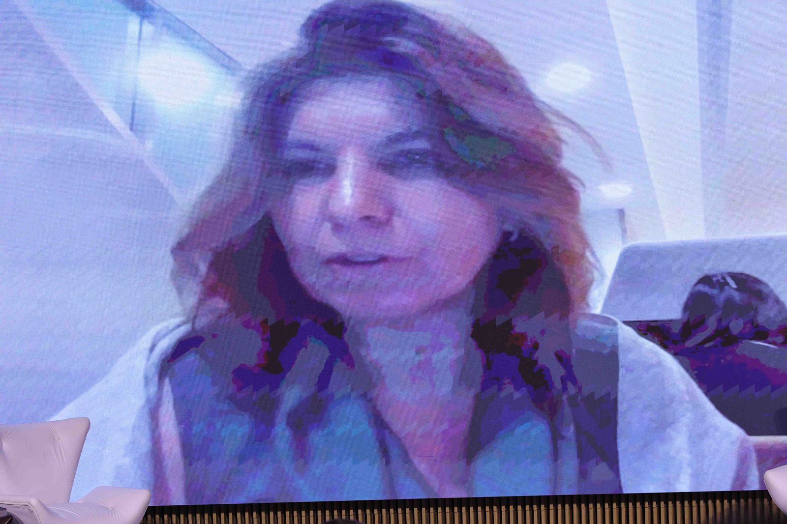 Uma captura de tela de videoconferência mostra a Dr.ª Teresa Cristina Cabral Santana (Ouvidoria da Secretaria-Geral da Presidência do TSE) em um grande monitor de LED no palco do III CIVIGE. Ela tem cabelos castanhos ondulados. Ela veste uma blusa escura e um casaco cinza claro. 