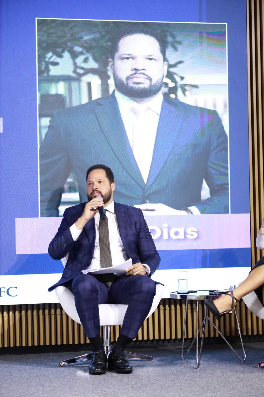 Dr. Tiago Dias da Silva, Juiz de Direito do TJCE, sentado em uma poltrona branca moderna em um palco durante uma conferência. Ele veste um terno cinza-chumbo, camisa social branca e uma gravata marrom com listras diagonais, segurando um microfone na mão direita. Atrás dele, há uma grande tela de LED.