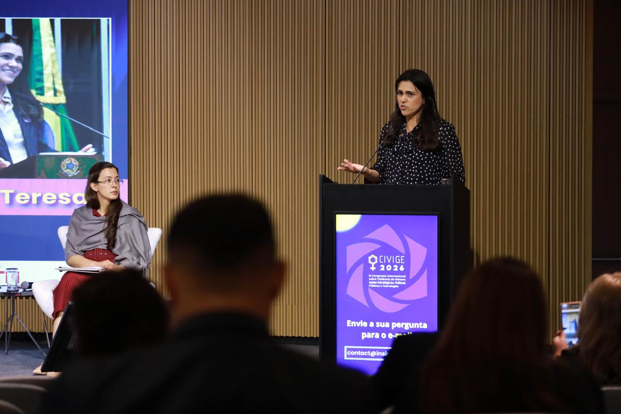 A Dr.ª Maria Teresa Prado, à direita, discursa em um púlpito preto durante o CIVIGE 2026, vestindo uma blusa preta com poás brancos. À esquerda, a Dr.ª Jaquelline Santos Silva observa sentada em uma poltrona branca, usando um xale cinza. Ao fundo, um painel de LED exibe uma transmissão de vídeo e o nome "Teresa", enquanto o monitor do púlpito mostra o logotipo do evento e instruções para envio de perguntas.