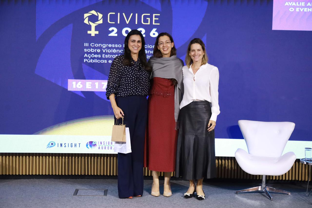 Três mulheres sorrindo em um palco de conferência, de pé em frente a uma grande tela de LED azul que exibe o logotipo "CIVIGE 2026" e o texto "III Congresso Internacional sobre Violência de Gênero". Da esquerda para a direita estão: Dr.ª Maria Teresa Prado, usando uma blusa preta de bolinhas brancas e calças escuras; Dr.ª Jaquelline Santos Silva, usando um vestido longo vermelho com um xale cinza; e Dr.ª Carla Pinheiro Bessa Von Bentzen Rodrigues, usando uma camisa branca e uma saia de couro preta. Uma poltrona branca moderna está à direita.