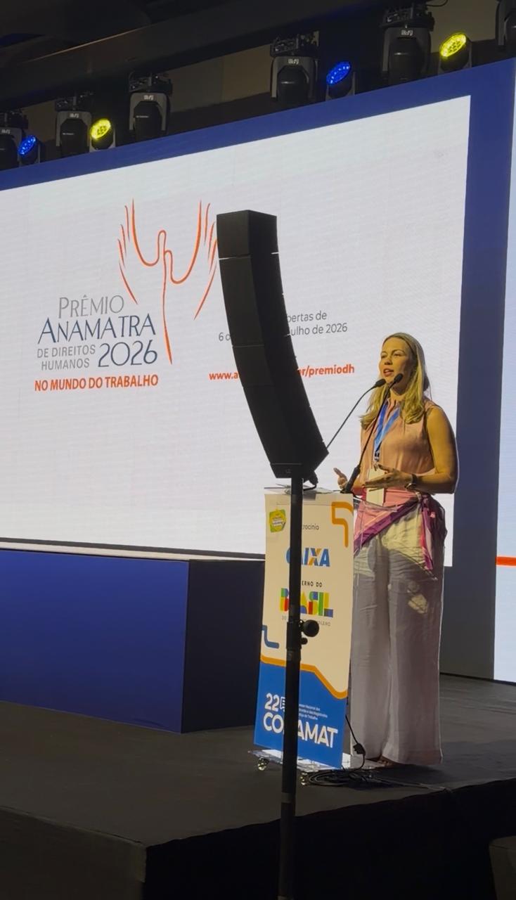 Uma mulher discursa em um púlpito. Ela veste uma blusa rosa e calça branca, posicionada à esquerda de um grande telão que exibe a identidade visual do congresso e informações sobre o Prêmio ANAMATRA. 