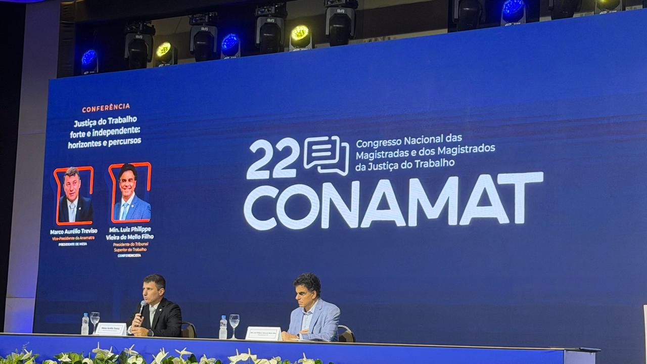 Dois palestrantes estão sentados à mesa de um painel durante o 22º CONAMAT. Ao fundo, um grande telão azul exibe o título da conferência "Justiça do Trabalho forte e independente: horizontes e percursos", acompanhado pelas fotos e nomes dos conferencistas. 