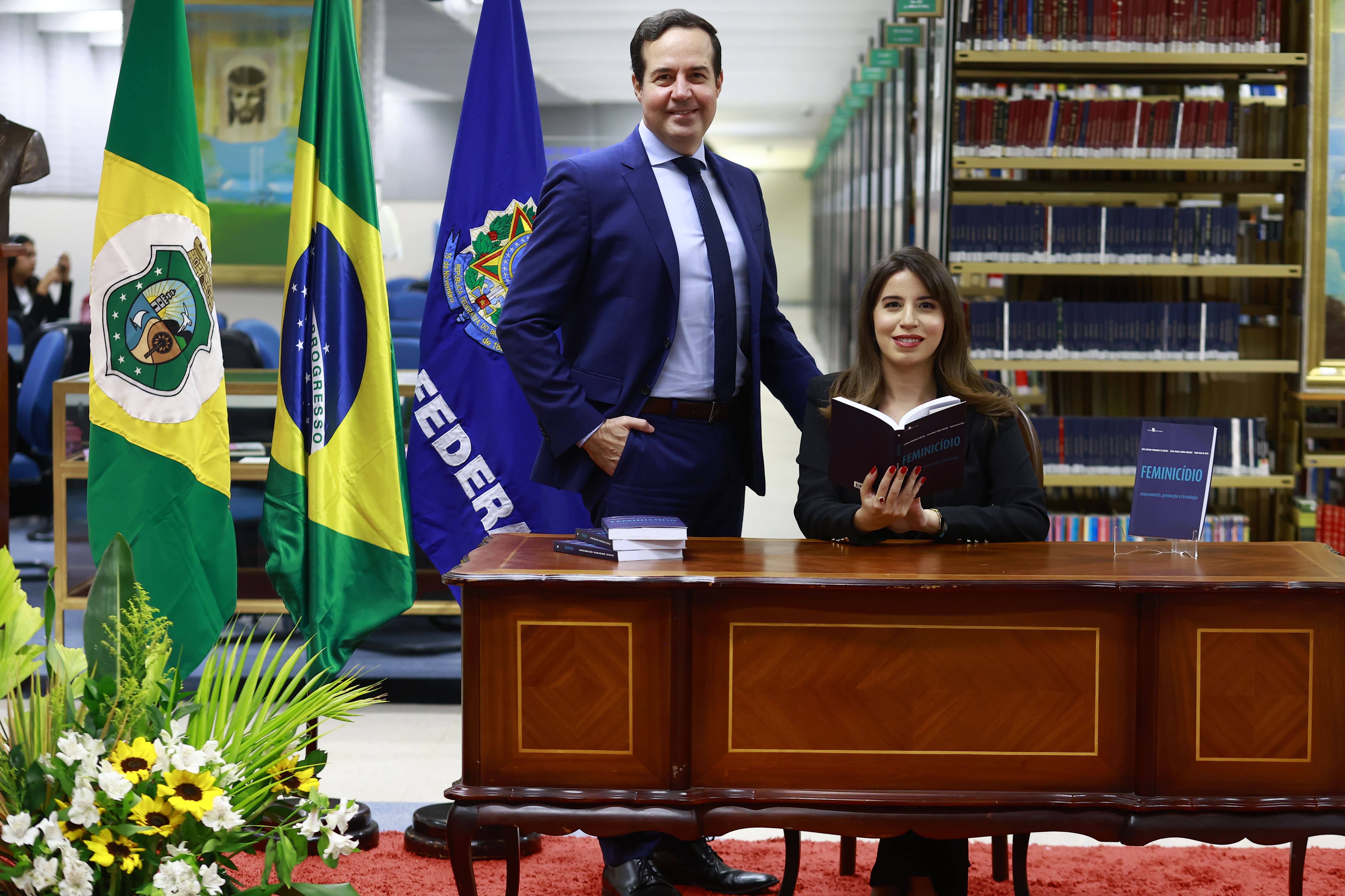 Um homem em pé e uma mulher sentada atrás de uma bancada de madeira em uma biblioteca. Eles sorriem para a câmera. A mulher segura um livro aberto intitulado "FEMINICÍDIO", e outro exemplar do mesmo livro está em destaque sobre a mesa. Ao fundo, estão posicionadas as bandeiras do Ceará, do Brasil e uma bandeira azul, além de estantes repletas de livros. Em primeiro plano, no canto inferior, há um arranjo de flores.