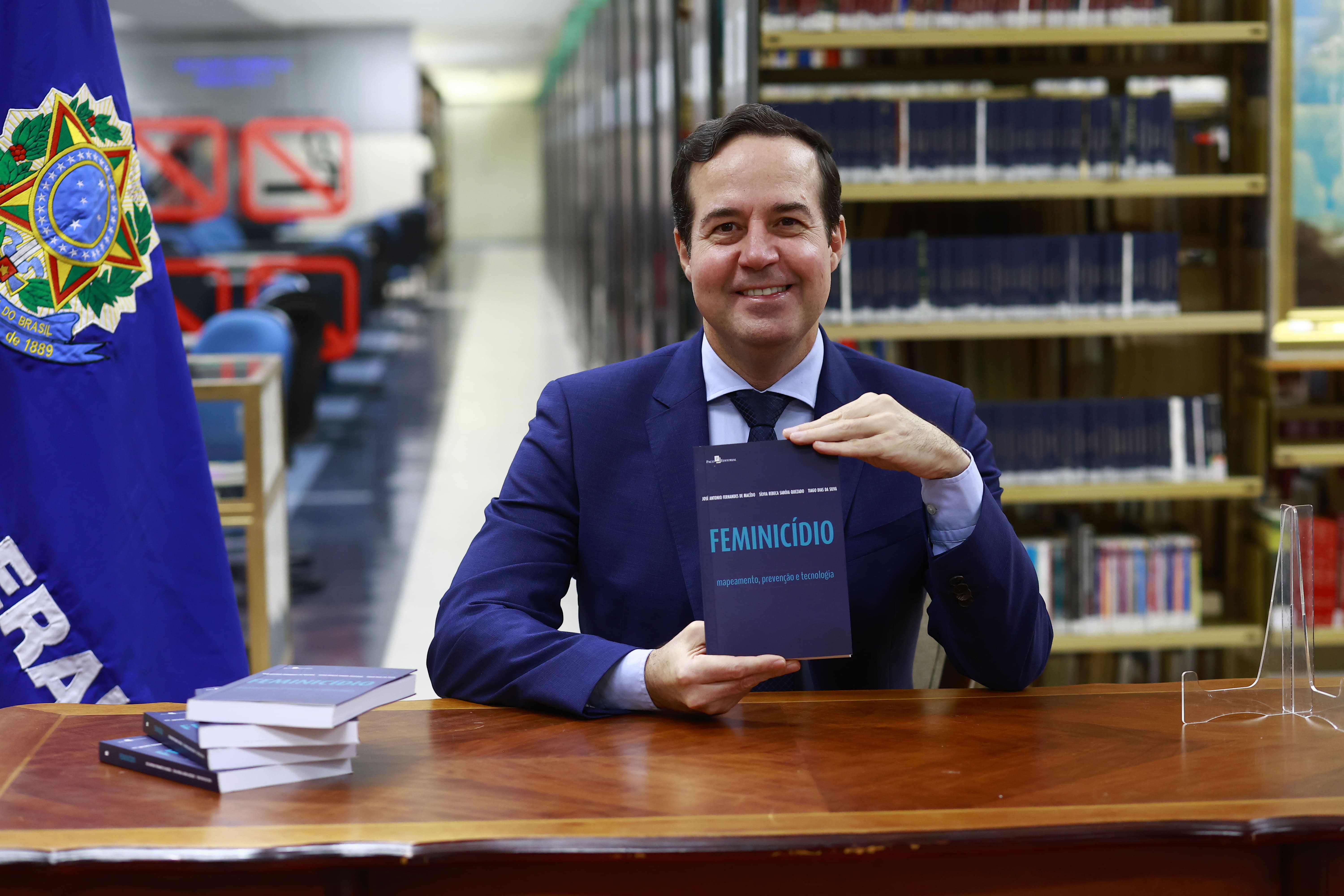 Um homem sorridente, vestindo paletó azul e gravata, sentado atrás de uma mesa de madeira. Ele segura à frente do corpo o livro intitulado "FEMINICÍDIO: mapeamento, prevenção e tecnologia". Sobre a mesa, há uma pequena pilha com outros exemplares do mesmo livro. Ao lado esquerdo, vê-se parte de uma bandeira azul com o brasão da República e a inscrição "FEDERAL". O fundo apresenta o corredor de uma biblioteca com estantes repletas de livros em perspectiva.