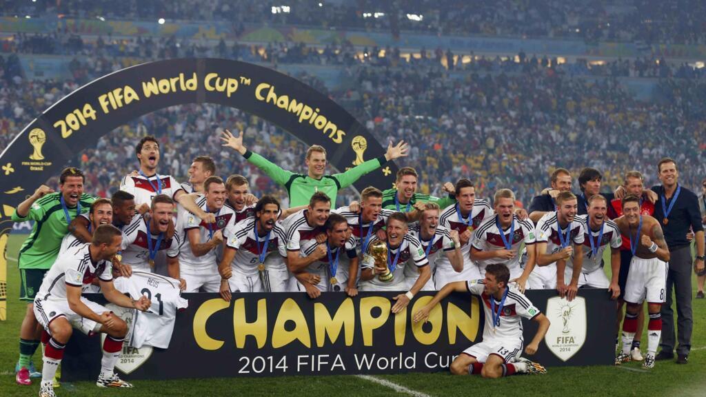 Jogadores da seleção alemã de futebol celebram no gramado de um estádio lotado. Eles estão reunidos atrás de uma estrutura preta com a palavra "CHAMPION" escrita em letras grandes e amarelas. Ao fundo, um arco preto traz a inscrição dourada "2014 FIFA World Cup Champions". No centro do grupo, os jogadores sorriem, se abraçam e usam medalhas de ouro no peito; um deles ergue a taça dourada da Copa do Mundo, enquanto o goleiro, vestindo uniforme verde, aparece no centro com os braços abertos. À direita, membros da comissão técnica de terno também comemoram.