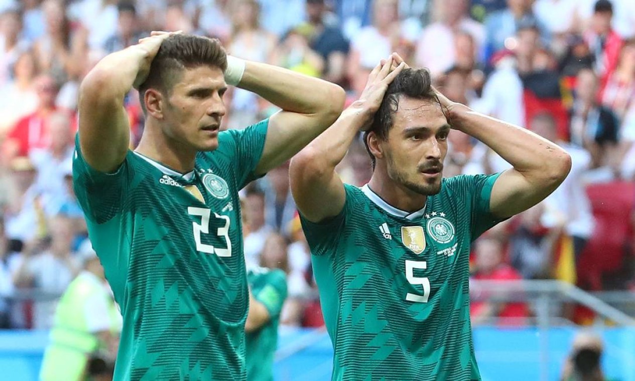 Dois jogadores da seleção alemã de futebol aparecem em primeiro plano com expressões de decepção e incredulidade. Ambos vestem o uniforme reserva verde, com detalhes geométricos texturizados e o escudo da federação alemã no peito. O jogador à esquerda veste o número 23 e o da direita veste o número 5; os dois estão com as mãos na cabeça e olhando fixamente para a frente. O fundo mostra as arquibancadas de um estádio de futebol com torcedores desfocados.