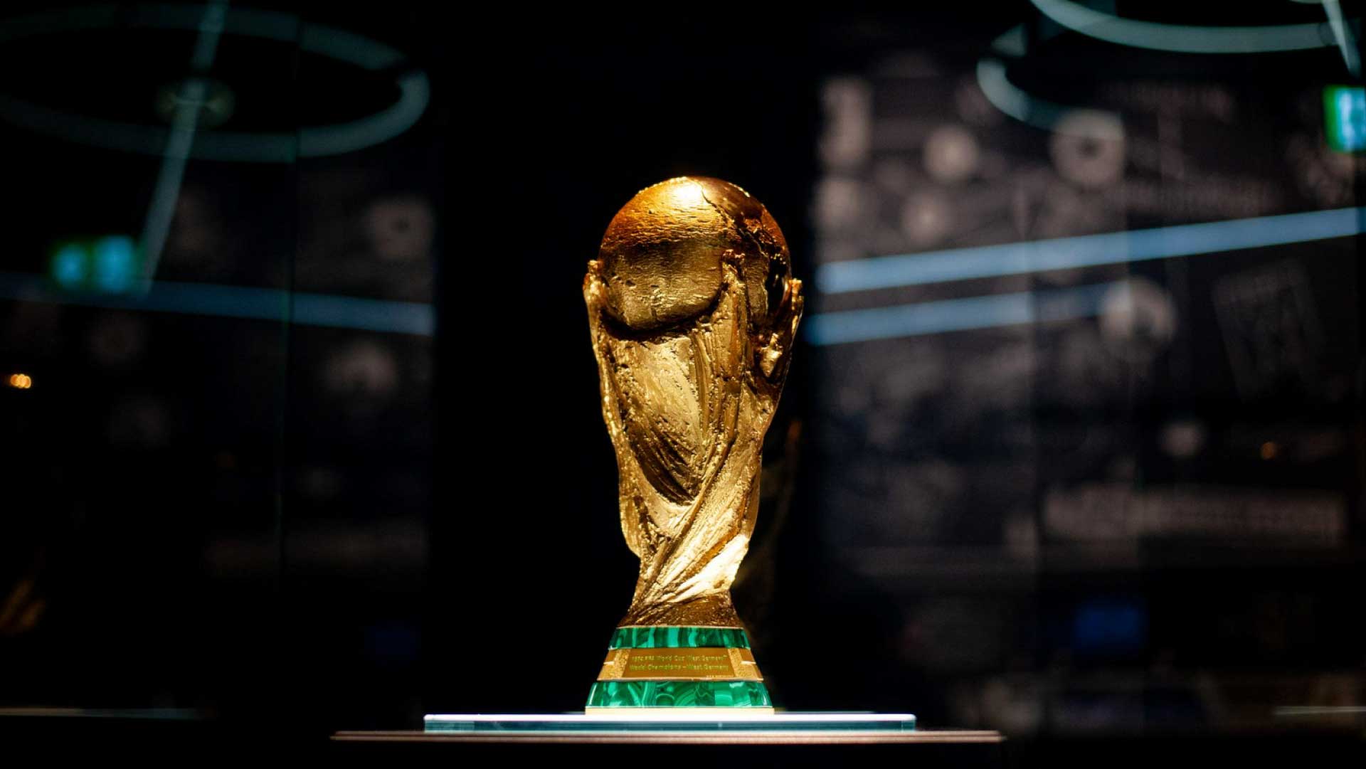 Fotografia em close-up do troféu original da Copa do Mundo da FIFA. A taça dourada está centralizada, iluminada por uma luz focada que destaca suas texturas e as duas figuras humanas estilizadas que sustentam o globo terrestre no topo. A base do troféu possui duas faixas verdes de mármore malaquita. O fundo é escuro e está completamente desfocado, exibindo sutilmente reflexos de luzes e estruturas de vidro de uma vitrine de exposição.
