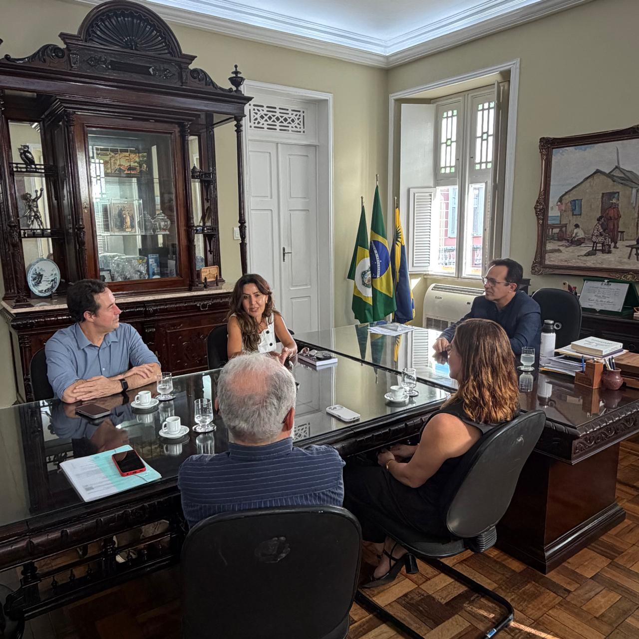Fotografia de uma reunião institucional no gabinete do reitor da Universidade Federal do Ceará. Representantes da UFC, do Insight Lab e da Procuradoria Regional do Trabalho estão sentados ao redor de uma grande mesa de vidro, em conversa sobre possibilidades de parceria. Ao fundo aparecem bandeiras, além de mobiliário de madeira, quadro, documentos e objetos de escritório.