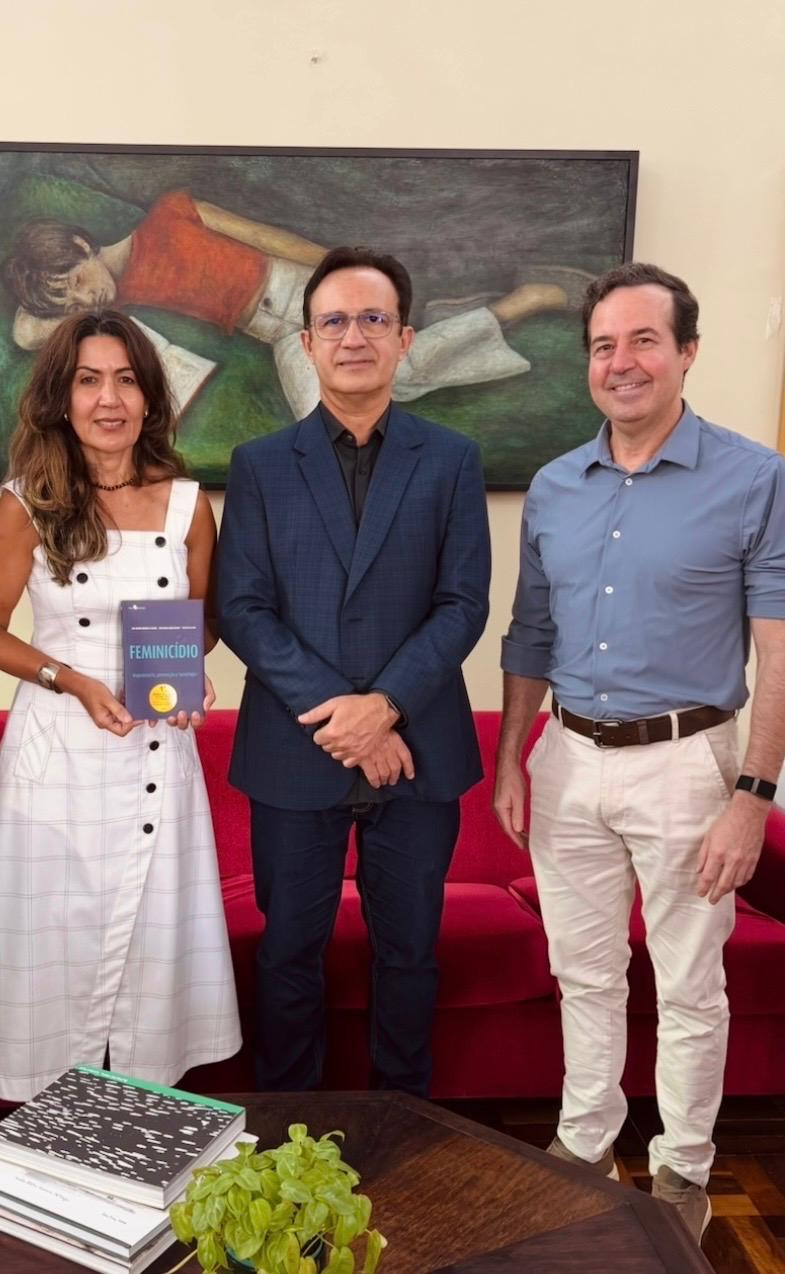 Fotografia de três pessoas em pé no gabinete da Reitoria da Universidade Federal do Ceará. À esquerda, a procuradora Ana Valéria Targino segura um exemplar do livro “Feminicídio - mapeamento, prevenção e tecnologia”. Ao centro está o reitor da UFC, Custódio Almeida, e à direita o professor José Macêdo, coordenador do Insight Data Science Lab. Ao fundo, há um sofá vermelho e uma pintura na parede; em primeiro plano, aparecem livros e uma planta sobre uma mesa.