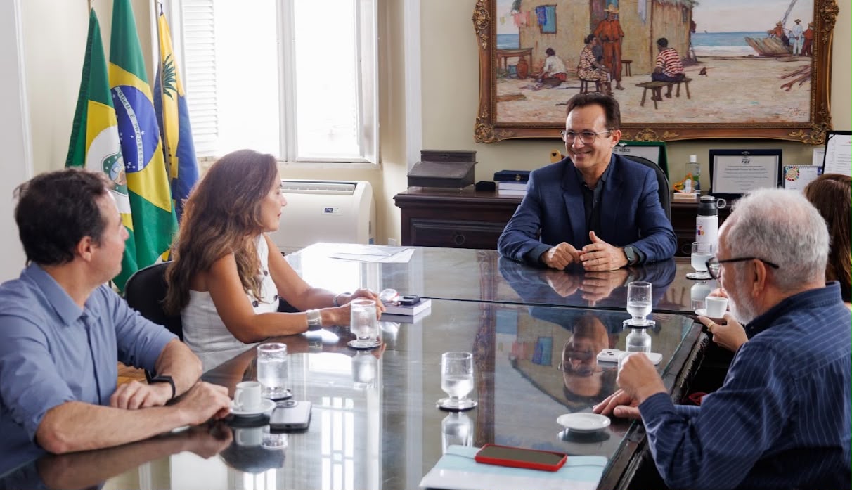 Fotografia de uma reunião institucional em uma sala da Universidade Federal do Ceará. Quatro pessoas aparecem sentadas ao redor de uma mesa de vidro, em conversa, enquanto outra pessoa é vista parcialmente ao fundo. Atrás delas estão bandeiras, além de um quadro e objetos de escritório. Sobre a mesa há copos de água, xícaras, celulares e documentos.